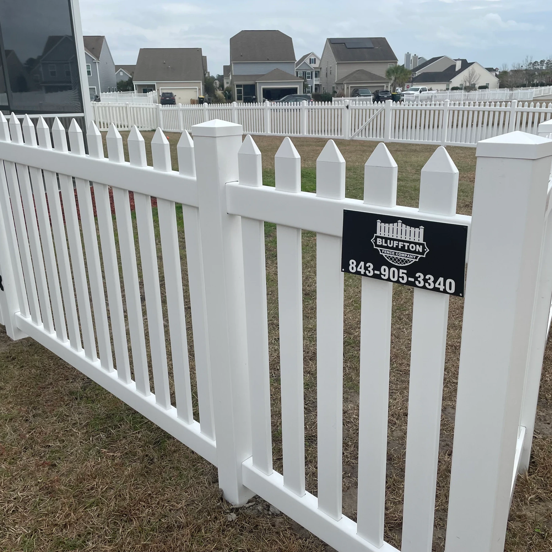 White vinyl picket fence with a Bluffton Fence & Gutter sign in a lakeside neighborhood